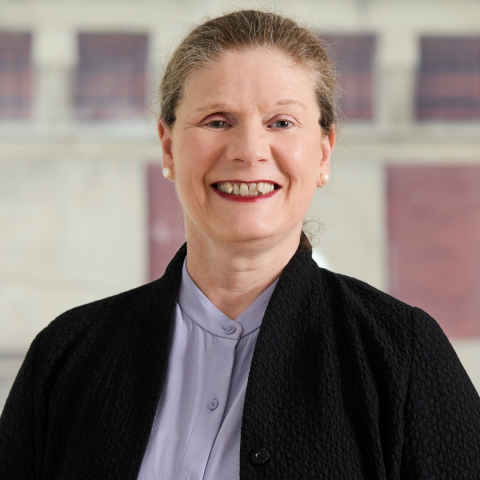 A professional portrait of a woman with brown hair pulled back, smiling and wearing a light purple button-down shirt under a black open jacket against a blurred background.