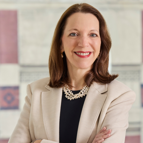 This image shows a woman posing for a professional headshot. She is smiling while wearing a beige blazer over a black top, complemented by a multi-stranded pearl necklace and small gold drop earrings. Her arms are loosely crossed in front of her.