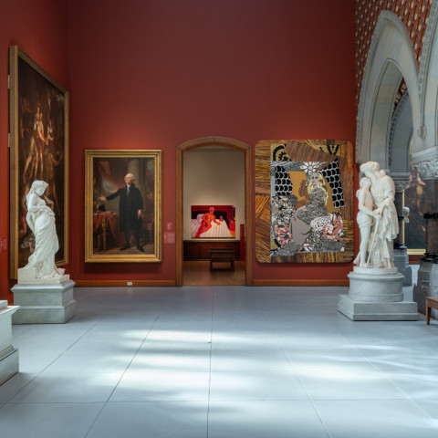 A museum gallery with deep red walls displays classical marble sculptures and framed oil paintings under gothic-style arches.
