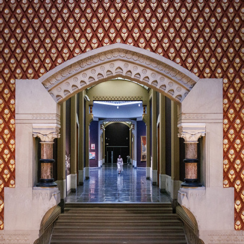 The image appears to show an interior hallway with patterned red and gold walls and a stone archway at the historic Pennsylvania Academy of the Fine Arts building in Philadelphia, designed by architect Frank Furness.