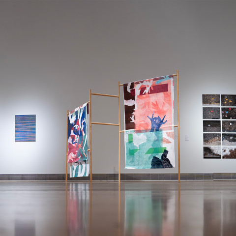 An installation view of a spacious gallery room showcases mixed-media contemporary art pieces, including colorful textile-like monoprints hanging on freestanding wooden quilt racks alongside grid-patterned photographic panels and textured abstract frames mounted on a minimalist white wall.