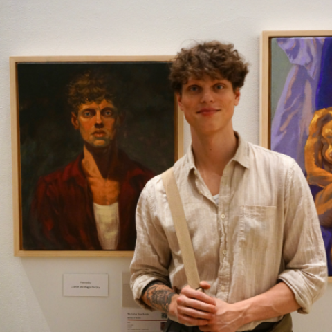 An artist with curly hair stands in a gallery next to his painted portrait.