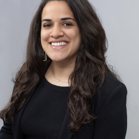 A smiling woman with long, wavy dark hair wears a black blazer and a delicate necklace against a plain grey background.