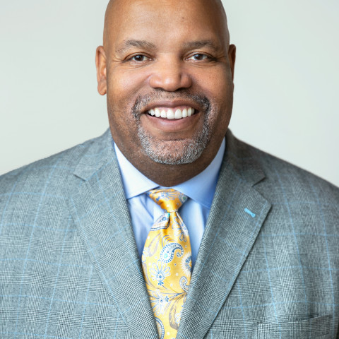 a professional headshot of a smiling, bald Black man with a neatly trimmed graying goatee and mustache, wearing a light gray patterned suit jacket, a light blue dress shirt, and a yellow paisley tie
