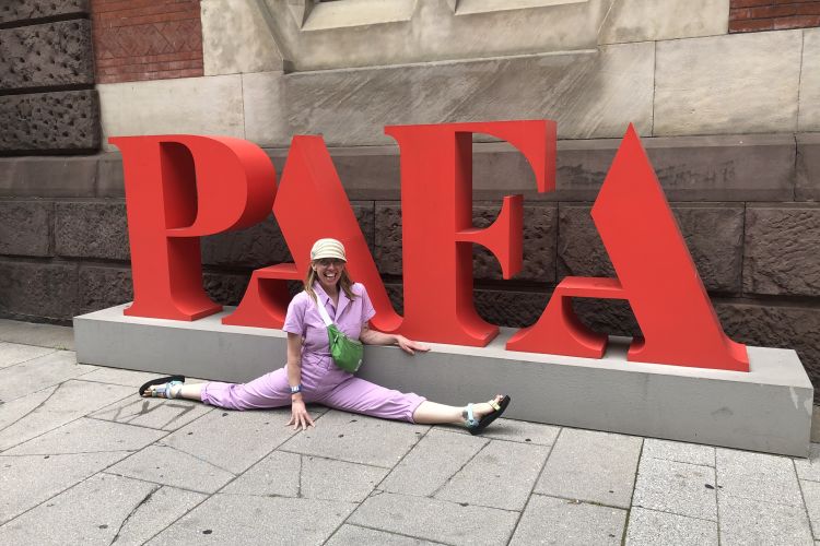 Kristy Jane smiles, doing the splits in front of a large PAFA sign