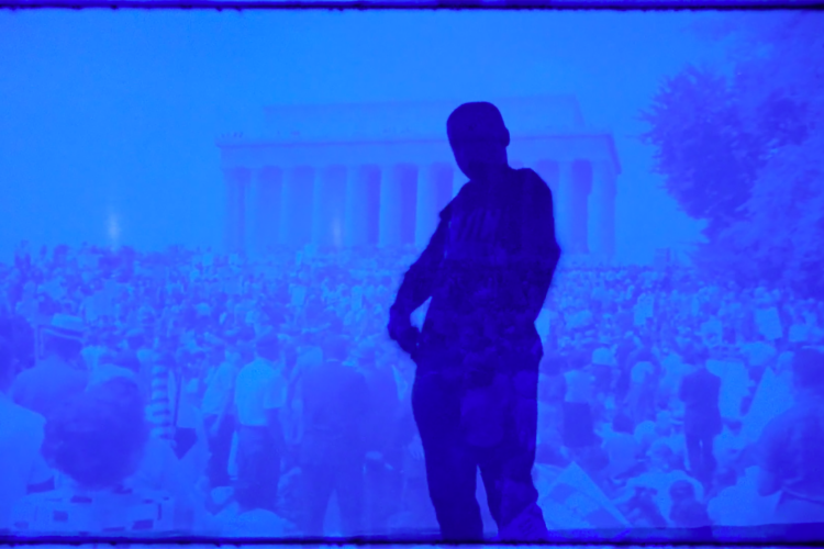 A blue-toned photograph of a figured shadow in front of a crowd at the Lincoln Memorial. 