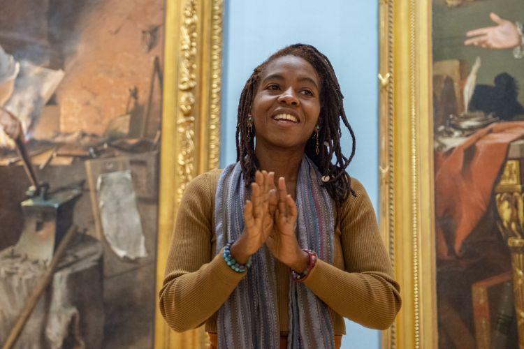 Photo of Misty Sol, a Black woman wearing a scarf, gesturing with the palms of her hands facing each other, and speaking with a smile. 