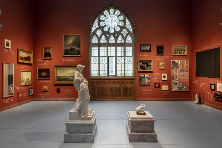 installation view of Lenka Clayton's work, "The True Story of a Stone". It is a gallery view with a classic Greek marble sculpture of a woman on the right and a small lifesize marble sculpture of a pekin duck. The walls have various types of paintings and ephemera, hung salon-style. An ornate rose-colored stained glass window is in the center of the gallery wall. 