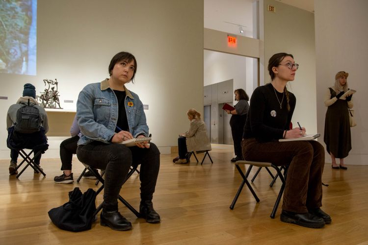 photo of four people sitting and two people standing in an art gallery sketching on small pads of paper. A small bronze sculpture is in the background. 