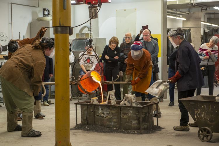 Photo of a a team of five people handling heavy equipment to pour molten bronze into molds. Behind the team stand five people looking intently at the pour in front of them. 