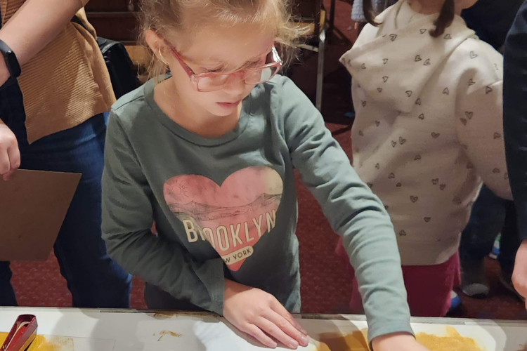 A child engaging in an art activity that involves sand and a roller. The person is wearing a long-sleeved shirt with the word "BROOKLYN" written across a heart graphic.