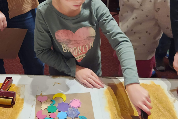 A child engaging in an art activity that involves sand and a roller. The person is wearing a long-sleeved shirt with the word "BROOKLYN" written across a heart graphic.