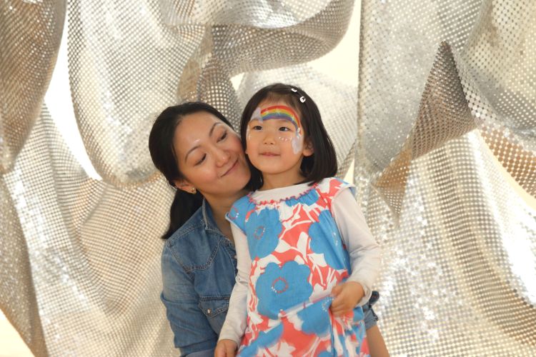 A mother and daughter stand smiling in front of a metallic backdrop, with the young girl sporting a rainbow painted on her forehead, suggesting they’re enjoying a fun face-painting event.