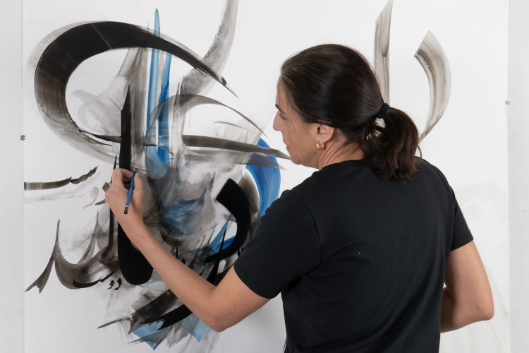 An artist with dark hair tied back in a ponytail and wearing a black t-shirt is captured from behind as she creates large-scale, expressive black and blue calligraphic strokes on a white canvas.