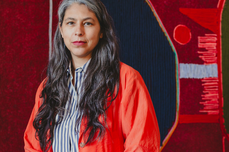 Artist Teresa Baker in a vibrant orange jacket and striped shirt sits in front of a red mixed-media artwork.