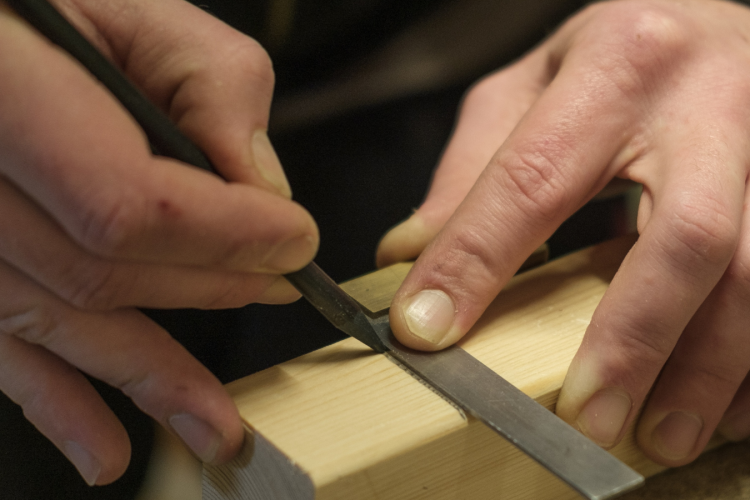  Visually searched image one sentence desciptive alt text A person's hands use a metal file to shape and smooth a precise notch in a light-colored wooden block.