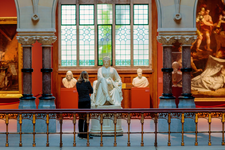 An art gallery visitor stands before a central white marble statue, flanked by busts and framed by ornate pillars and a large stained-glass window.