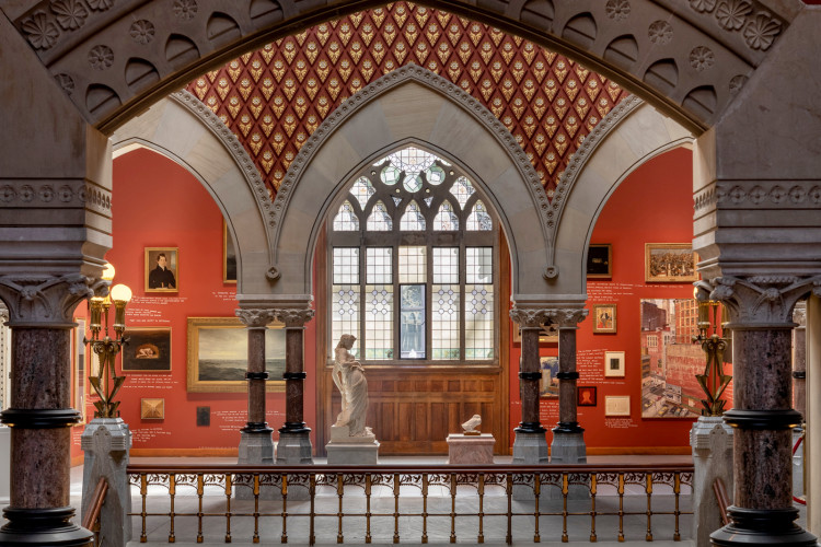 Gothic-style gallery with arched windows, ornate red and gold ceiling, and classical sculptures. Warm lighting and red walls create an elegant ambiance.