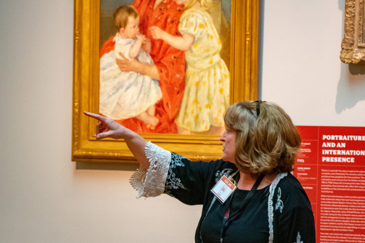 A woman in a black dress with lace details gestures towards a painting of a woman and two children. She holds a book, conveying an educational setting.