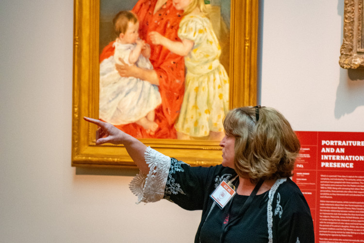 A woman in a black dress with lace details gestures towards a painting of a woman and two children. She holds a book, conveying an educational setting.