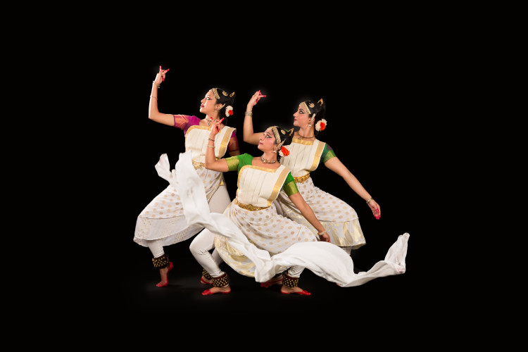 three Indian classical dancers in white and gold sarees perform Mohiniyattam with graceful poses and flowing fabrics against a solid black background.