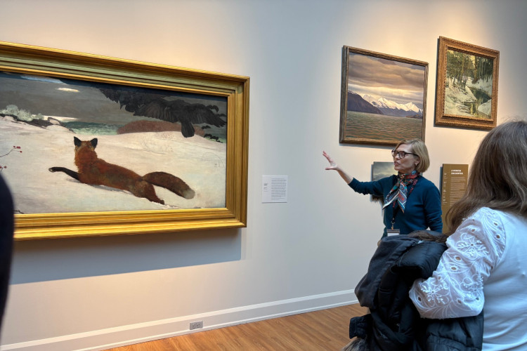 A museum guide explains a painting of a fox in snow to an engaged visitor. The artwork is large, set in a gold frame, with serene mountain scenes nearby.