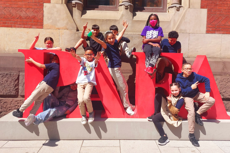 A group of smiling children pose playfully on large red "PAFA" letters against a brick building. Bright, cheerful atmosphere with kids showing excitement.