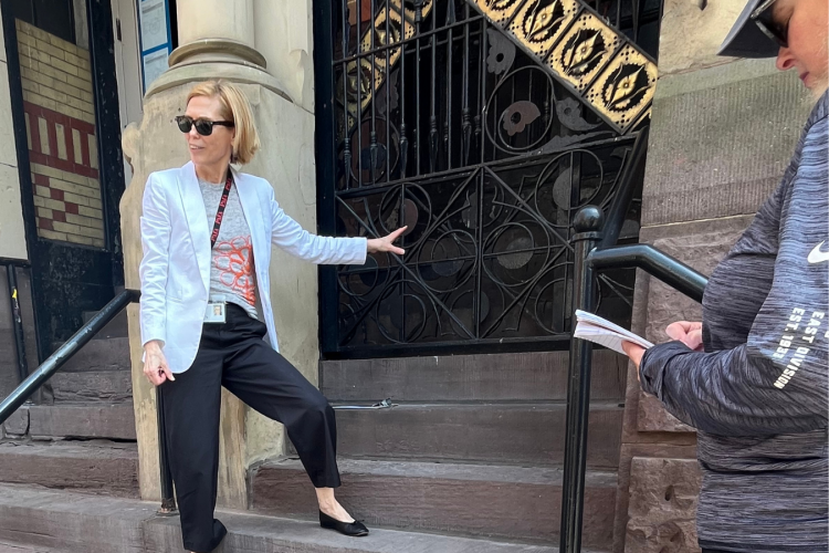 A woman with blonde hair, wearing a white blazer, dark pants, and sunglasses, stands on concrete steps while gesturing toward a decorative black iron gate.