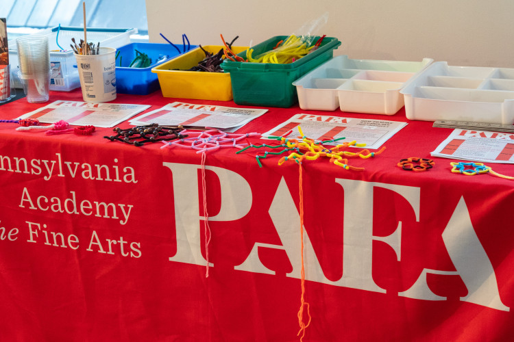 A table at the Pennsylvania Academy of the Fine Arts, covered with a red cloth displaying its name. Art supplies and handouts are neatly arranged on top.