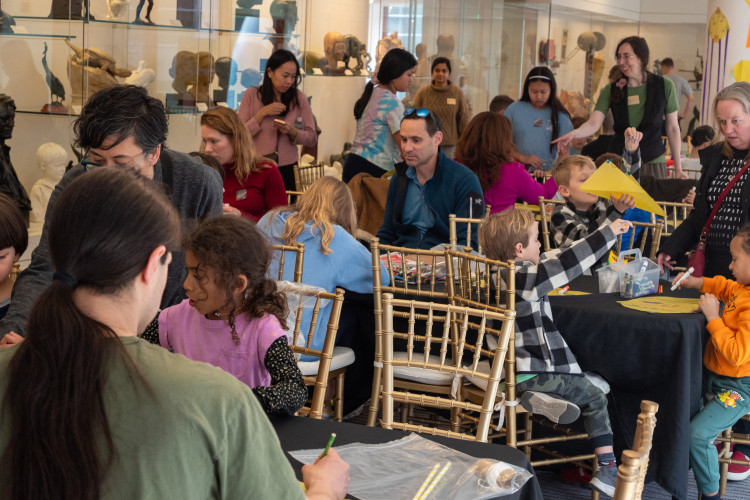 A lively room with children and adults engaged in arts and crafts at a museum. The scene is busy, with sculptures displayed in the background.