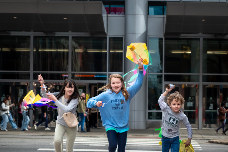 Three children joyfully run with kites on a wooden walkway. They wear colorful clothing and smile excitedly, with a busy city street in the background.