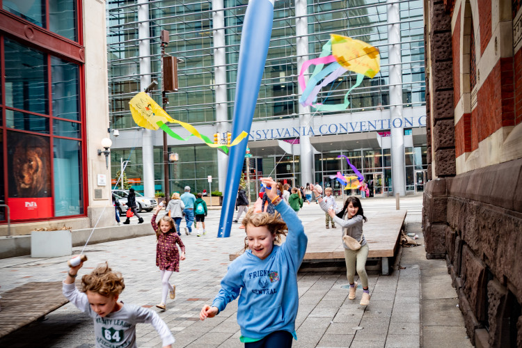 Children joyfully run with colorful streamers near a giant paintbrush sculpture outside the Pennsylvania Convention Center, exuding excitement and playfulness.