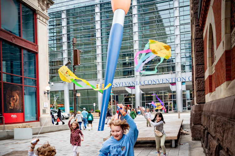 Children joyfully run with colorful streamers in front of the Pennsylvania Convention Center. A giant paintbrush sculpture adds a whimsical touch.