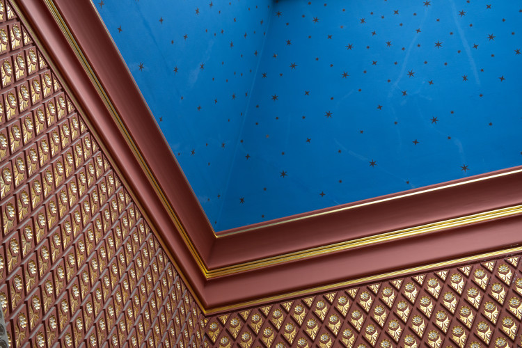 A high-angle shot captures an ornate corner ceiling featuring a bright blue panel dotted with small gold stars, framed by thick reddish-brown and gold trim above walls covered in geometric gold and brown patterns.