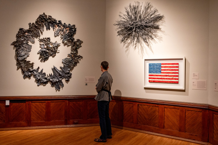 A person in an art gallery stands looking at a large, circular textured sculpture on the wall, with a spiky burst sculpture and an encaustic painting of the American flag displayed nearby.