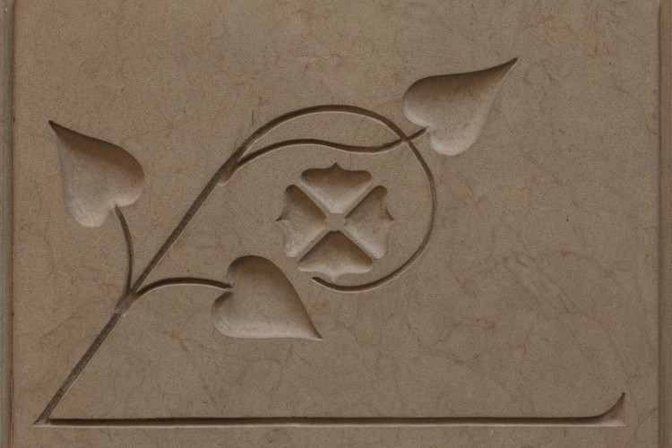 A rectangular stone panel features a low-relief architectural carving of a stylized vine with three heart-shaped leaves curving gracefully around a central quatrefoil flower emblem.