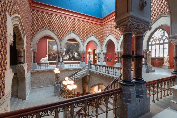 An ornate, Gothic Revival museum interior features patterned red walls, pointed stone arches, a bright blue ceiling, and a grand central staircase illuminated by spherical lamps.