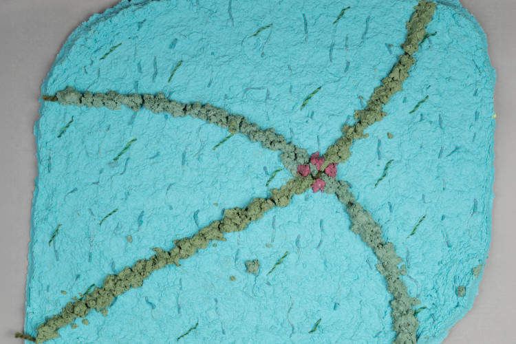 A textured turquoise handmade paper artwork features intersecting grayish-green fibrous lines that meet at a central cluster of small pink floral shapes.