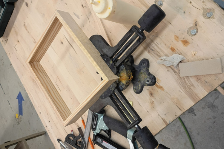a wooden picture frame corner being assembled and held at a 90-degree angle using a cast iron miter clamp or corner vise mounted to a workbench. 