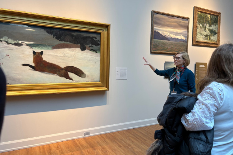 A museum guide explains a painting of a fox in snow to an engaged visitor. The artwork is large, set in a gold frame, with serene mountain scenes nearby.