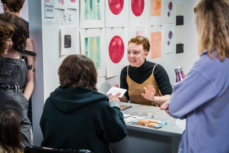 An art student with short hair smiles and speaks with visitors gathered around a workspace table inside a PAFA studio during an open studio night.